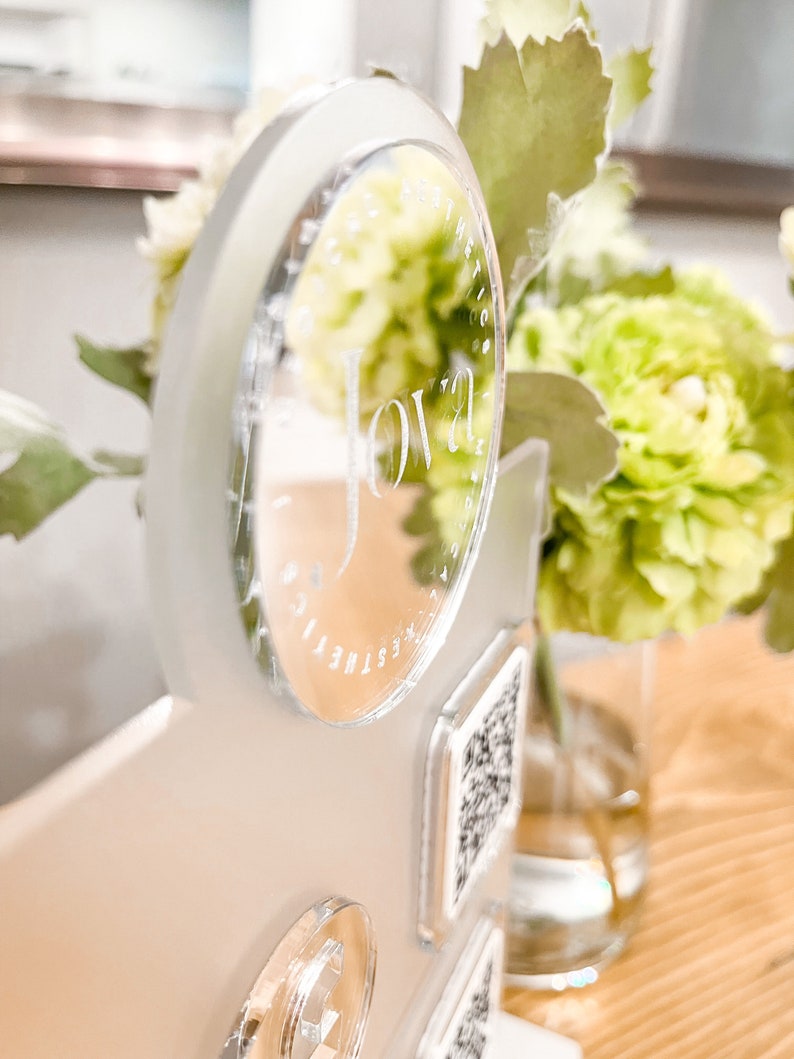 May include: A clear acrylic sign with the word "Jova" etched into it. The sign is attached to a white surface with a clear acrylic circle and a QR code.