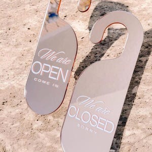 以下が含まれることがあります： 2つのローズゴールドのドアハンガー。1つは白い筆記体で「We are OPEN, COME IN」と書かれています。もう1つは白い筆記体で「We are CLOSED, SORRY」と書かれています。ハンガーは吊り下げ用の湾曲した上部が付いています。