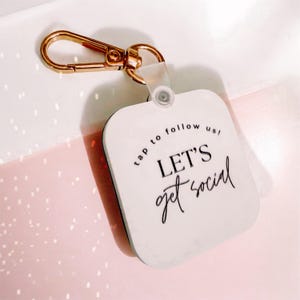 Może przedstawiać: Brelok w kształcie białego kwadratu z napisem "LET'S get social" czcionką. Brelok ma złoty karabińczyk i przezroczystą plastikową pętlę. Słowa "tap to follow us!" są wydrukowane nad głównym tekstem.
