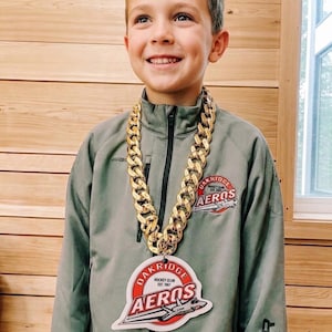May include: A large gold-coloured plastic chain necklace featuring a circular pendant with a red and white logo that reads Oakridge Hockey Club Est. 1967 Aeros, worn over a grey zip-up jacket with a matching team logo on the chest.
