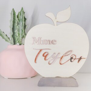 May include: A white wooden apple-shaped sign with a gray stand. The sign has the text "Mme Taylor" written in rose gold lettering.