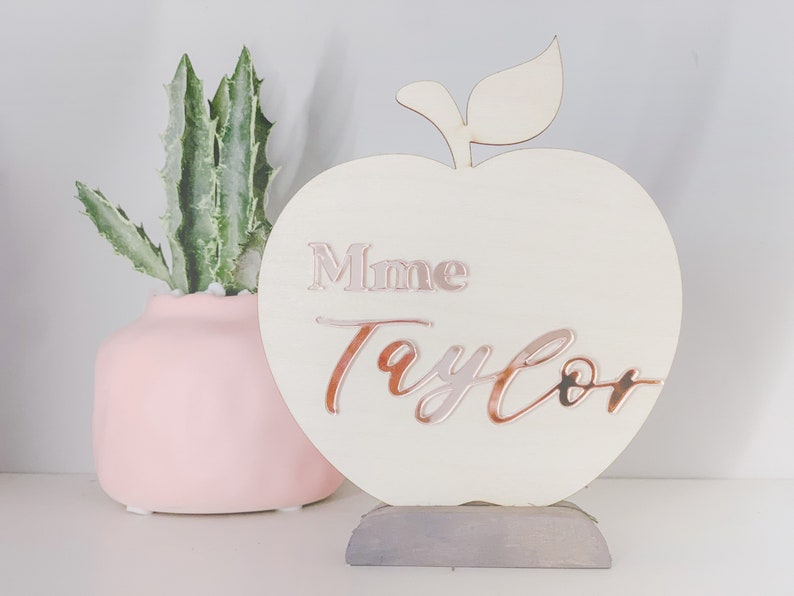 May include: A white wooden apple-shaped sign with a gray stand. The sign has the text "Mme Taylor" written in rose gold lettering.