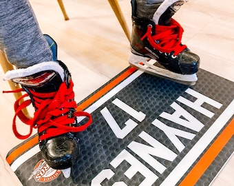 May include: A black and orange hockey mat with the words "SENATE 12" printed in white. A person is wearing black and red ice skates on the mat.