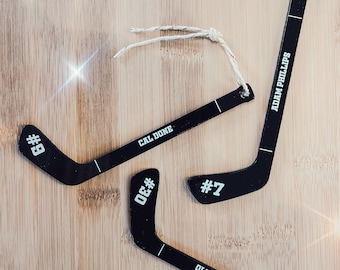 Könnte beinhalten: Drei schwarze Hockey-Schläger-Anhänger mit weißem Band zum Aufhängen. Die Schläger sind mit Namen und Nummern personalisiert: Cal Done #9, Oliver Hibbits #7 und Adam Phillips #5.
