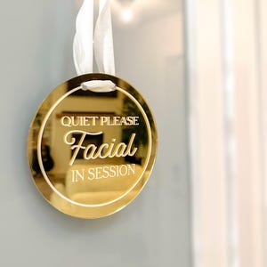May include: A circular gold-toned reflective sign hanging from a white ribbon. The sign features the white text "QUIET PLEASE" at the top, "Facial" in a larger script font in the centre, and "IN SESSION" at the bottom, all with a white border.