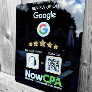 May include: A black rectangular sign with white and green text that says "REVIEW US ON Google" with the Google logo. It also says "TAP YOUR PHONE" and "SCAN QR" with a QR code and a phone icon. The sign also says "NowCPA Chartered Professional Accountants".