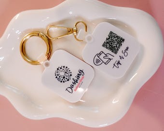 Könnte beinhalten: Zwei weiße quadratische Schlüsselanhänger mit goldenen geteilten Ringen. Ein Schlüsselanhänger hat ein schwarz-weißes Logo mit dem Text "Dashing Bubble Tea Shop" und eine schwarz-weiße Grafik eines Ballons. Der andere Schlüsselanhänger hat einen schwarz-weißen QR-Code und den Text "Tap or Scan".