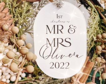 Pode incluir: Ornamento redondo branco com o texto "1º Natal como MR & MRS Olivera 2022" pendurado em uma guirlanda de Natal com paus de canela e agulhas de pinho.