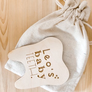 May include: A wooden tooth-shaped baby teether with the text "Leo's baby teeth" engraved on it. The teether is in a white cloth bag.