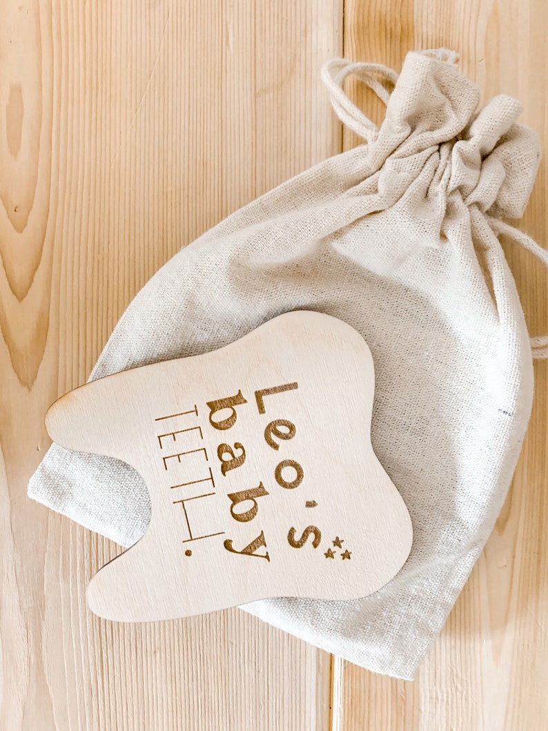 May include: A wooden tooth-shaped baby teether with the text "Leo's baby teeth" engraved on it. The teether is in a white cloth bag.