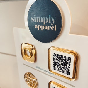May include: A white display board with a black circle featuring the text "simply apparel". The board has several gold-colored square and round pins with QR codes and other designs. The text "@hellosimplyapparel" is printed on the board.