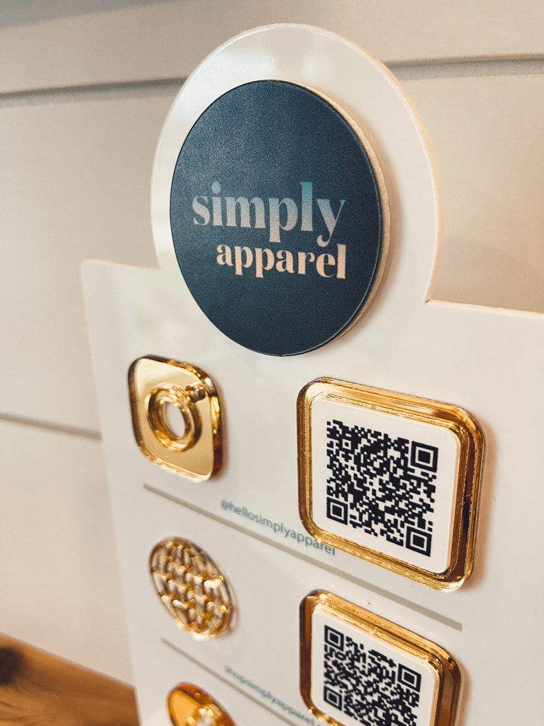 May include: A white display board with a black circle featuring the text "simply apparel". The board has several gold-colored square and round pins with QR codes and other designs. The text "@hellosimplyapparel" is printed on the board.