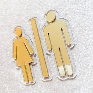 Peut inclure: Un panneau de toilettes de couleur or avec un symbole masculin et féminin. Les symboles sont entourés de plastique transparent.