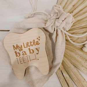 May include: A wooden tooth-shaped keepsake box with the text "My Little baby TEETH!" engraved on it. The box is in a small, beige drawstring bag.