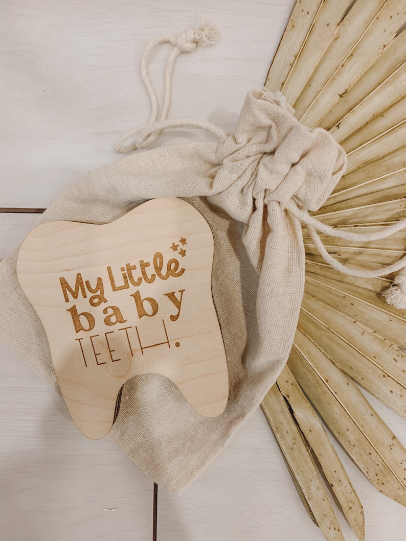 May include: A wooden tooth-shaped keepsake box with the text "My Little baby TEETH!" engraved on it. The box is in a small, beige drawstring bag.