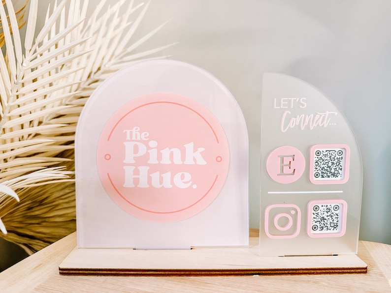 Peut inclure: Une enseigne en acrylique blanche avec un cercle rose qui dit "The Pink Hue." L'enseigne a une base en bois. &Agrave; c&ocirc;t&eacute; se trouve une enseigne en acrylique blanche avec le texte "Let's Connect..." et des ic&ocirc;nes pour Instagram, l'e-mail et un code QR.