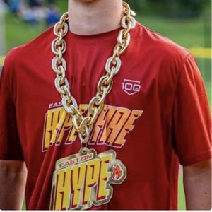 Peut inclure: Un t-shirt rouge à manches courtes avec le texte jaune et blanc Easton Hype et un logo 100 ans. Un grand collier à chaîne en plastique doré épais avec un pendentif Easton Hype assorti est porté autour du cou sur le t-shirt.