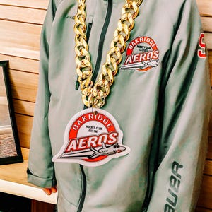 Peut inclure: Un enfant portant une veste grise avec un collier de chaîne dorée et un autocollant blanc avec le texte "Oakridge Hockey Club Est. 1987 Aeros" et un avion de dessin animé.