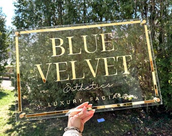 May include: A clear acrylic sign with gold trim and gold lettering. The sign reads "BLUE VELVET Esthetics LUXURY FACIALS". The sign is held up by a person's hand in front of a green background.
