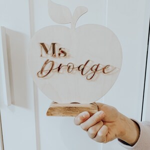 May include: Wooden apple-shaped teacher desk sign with a stand. The sign says "Ms. Drodge" in gold lettering.