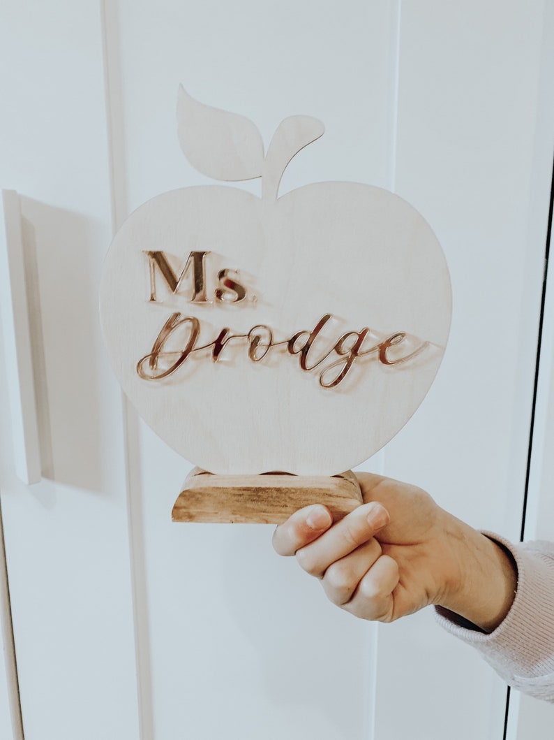 May include: Wooden apple-shaped teacher desk sign with a stand. The sign says "Ms. Drodge" in gold lettering.