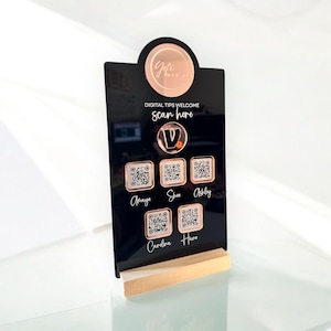 May include: A black acrylic digital tips welcome sign with a rose gold logo and QR codes. The sign reads "DIGITAL TIPS WELCOME scan here" and lists names below the QR codes. The sign is supported by a light wood base.