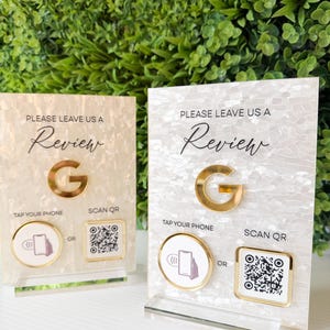 May include: Two pearlescent review stands with gold detailing. Each stand displays "PLEASE LEAVE US A Review" and a gold "G" logo. Options to "TAP YOUR PHONE" or "SCAN QR" are below for reviews. The stands are on a clear base.