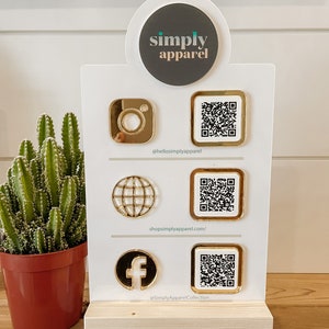 May include: A white sign with the words "simply apparel" in a circle at the top. Below the circle are three rows of gold icons and QR codes. The first row has an Instagram icon and a QR code with the text "@hellosimplyapparel". The second row has a globe icon and a QR code with the text "shopsimplyapparel.com/". The third row has a Facebook icon and a QR code with the text "@SimplyApparelCollection".