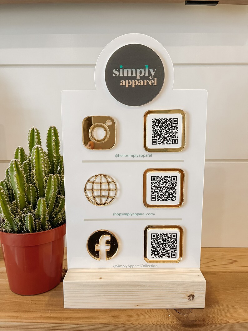 May include: A white sign with the words "simply apparel" in a circle at the top. Below the circle are three rows of gold icons and QR codes. The first row has an Instagram icon and a QR code with the text "@hellosimplyapparel". The second row has a globe icon and a QR code with the text "shopsimplyapparel.com/". The third row has a Facebook icon and a QR code with the text "@SimplyApparelCollection".