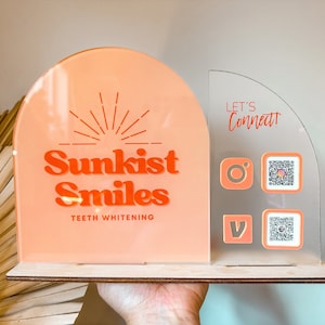 Peut inclure: Une enseigne en acrylique rose et blanche avec le texte "Sunkist Smiles Teeth Whitening" et un graphique de soleil. L'enseigne est sur une base en bois. Une enseigne en acrylique transparente avec le texte "Let's Connect!" et des ic&ocirc;nes de m&eacute;dias sociaux et des codes QR est fix&eacute;e sur le c&ocirc;t&eacute; droit de l'enseigne.