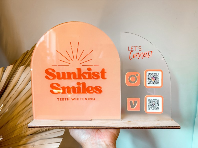Peut inclure: Une enseigne en acrylique rose et blanche avec le texte "Sunkist Smiles Teeth Whitening" et un graphique de soleil. L'enseigne est sur une base en bois. Une enseigne en acrylique transparente avec le texte "Let's Connect!" et des ic&ocirc;nes de m&eacute;dias sociaux et des codes QR est fix&eacute;e sur le c&ocirc;t&eacute; droit de l'enseigne.