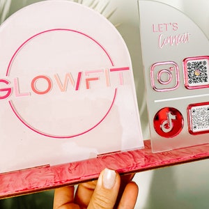 Peut inclure: Une enseigne en acrylique rose et blanche avec le texte "GLOWFIT" dans un cercle. L'enseigne a une base en marbre rose. Le c&ocirc;t&eacute; droit de l'enseigne porte le texte "LET'S Connect" et des ic&ocirc;nes pour Instagram, TikTok et des codes QR.