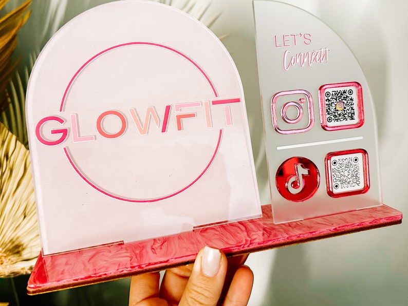 Peut inclure: Une enseigne en acrylique rose et blanche avec le texte "GLOWFIT" dans un cercle. L'enseigne a une base en marbre rose. Le c&ocirc;t&eacute; droit de l'enseigne porte le texte "LET'S Connect" et des ic&ocirc;nes pour Instagram, TikTok et des codes QR.