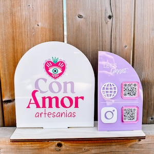 Peut inclure: Une enseigne blanche et violette avec le texte "Con Amor artesan&iacute;as" et un c&oelig;ur rose avec un motif d'&oelig;il. Une enseigne violette &agrave; c&ocirc;t&eacute; dit "Let's Connect" et a un globe, Instagram et deux codes QR.