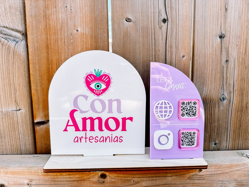 Peut inclure: Une enseigne blanche et violette avec le texte "Con Amor artesan&iacute;as" et un c&oelig;ur rose avec un motif d'&oelig;il. Une enseigne violette &agrave; c&ocirc;t&eacute; dit "Let's Connect" et a un globe, Instagram et deux codes QR.