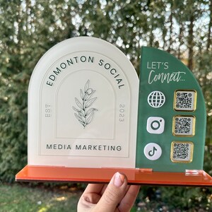 Peut inclure: Panneau en acrylique blanc et vert avec le texte "Edmonton Social" et "EST 2023" et "Media Marketing". Le panneau comporte &eacute;galement un petit motif de branche d'olivier. Sur le c&ocirc;t&eacute; droit du panneau se trouve un panneau en acrylique vert avec le texte "Let's Connect..." et quatre carr&eacute;s blancs avec des ic&ocirc;nes et des codes QR.