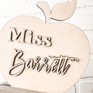 May include: A wooden apple with a 3D name cutout that reads "Miss Barrett". The apple is on a wood base. The text "Wood base + 3D name" is below the apple. The hashtag "#teacherappreciationweek" is below the text.