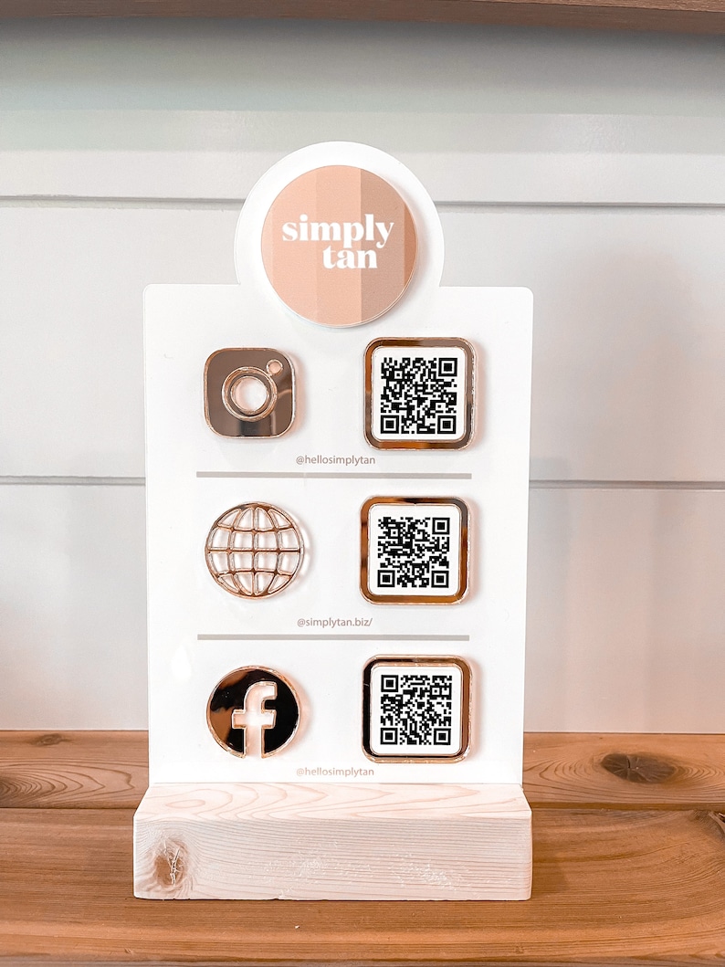 May include: A white sign with a pink circle that says "simply tan" on it. The sign has three social media icons with QR codes next to them. The icons are Instagram, a globe, and Facebook. The sign is on a wooden surface.