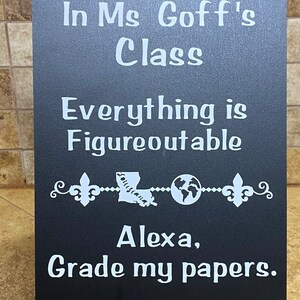 May include: A black chalkboard sign with white lettering. The sign reads "In Ms. Goff's Class, Everything is Figureoutable, Alexa, Grade my papers." Decorative elements include fleur-de-lis and a map of Louisiana.