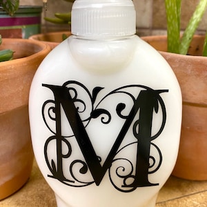 May include: White ceramic soap dispenser with a black letter "M" and decorative swirls. The dispenser has a pump top.