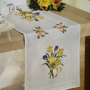 May include: A white rectangular table runner featuring embroidered floral motifs of yellow and purple daffodils tied with yellow ribbons, along with smaller orange flower clusters. The fabric has a textured border detail along the long edges.