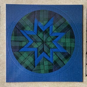 May include: A square blue card features a circular design with a starburst pattern. The star is made of blue ribbon and the background is a green and blue plaid. The card has a textured surface.