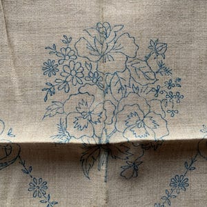 Op de afbeelding: Een beige linnen stof met blauwe geborduurde bloemenmotieven. Het ontwerp toont een boeket bloemen met bladeren en stelen.