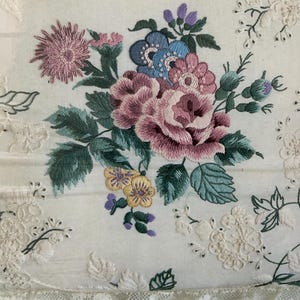 Könnte beinhalten: Gesticktes Blumendesign mit einem Bouquet aus rosa, lila, blauen und gelben Blumen mit grünen Blättern. Die Stickerei befindet sich auf einem weißen Stoff mit Spitzenbesatz und zusätzlichen gestickten floralen Akzenten.