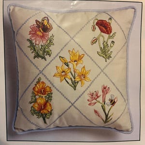 以下が含まれることがあります： 刺繍された花の模様が施された正方形の装飾用枕。クリーム色の枕は、青色のステッチの格子模様が特徴です。各正方形には、蝶、ポピー、ユリなど、さまざまな花が含まれています。青い縁取り。