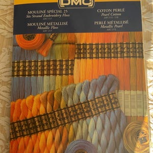May include: A close-up of a DMC embroidery floss color card. The card features a variety of colors, including orange, yellow, blue, and green. The card also includes the text "Mouline Special 25", "Six Strand Embroidery Floss", "Art 117", "Mouline Metallise", "Metallic Floss", "Art 317", "Coton Perle", "Pearl Cotton", "Art 115-116", "Perle Metallise", "Metallic Pearl", and "Art 315".