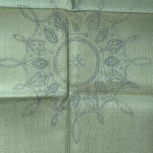 May include: A light green square textile with a detailed, symmetrical design in dark blue. The pattern features a central circle with a floral element, surrounded by stylized leaves and interconnected lines, creating a mandala-like appearance.
