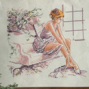 May include: A cross-stitch embroidery on white fabric depicting a person with auburn hair wearing a lavender dress, sitting and tying ballet slippers. To the left, a cluster of purple flowers is visible, and a grid pattern appears on the right.