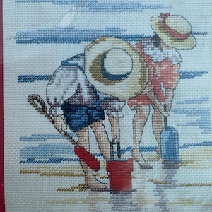 Puede incluir: Patrón de punto de cruz que representa dos figuras en una playa, con sombreros y pantalones cortos, con un cubo rojo y una botella azul. Las figuras están agachadas, posiblemente recogiendo algo. El fondo muestra un cielo y agua.
