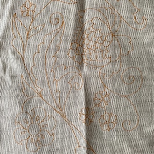 May include: A piece of light beige, textured fabric featuring a printed orange outline of a floral design. The pattern consists of large, stylized leaves, swirling vines, and several small blossoms arranged in a flowing, organic composition.